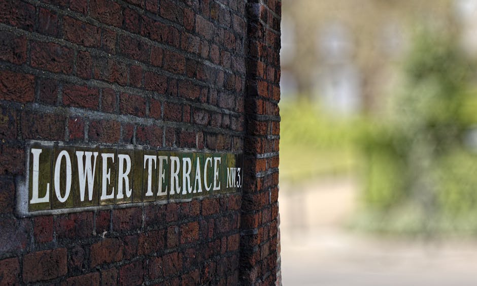 Close-up view of a brick wall featuring a rectangular ceramic street sign with the inscription 'LOWER TERRACE NW3' in white letters on a dark background, mounted securely on the wall. The textured red