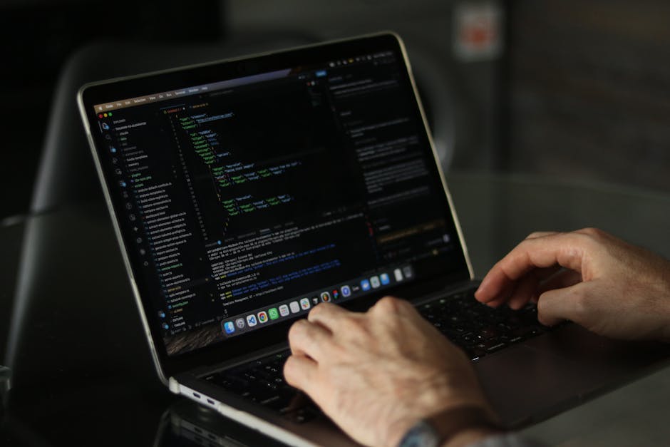 A close-up of a person's hands typing on a MacBook laptop placed on a dark surface. The laptop screen displays lines of coding or programming software, with visible text in various colors including gr